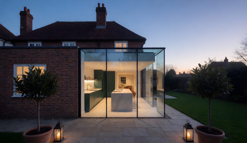 Frameless glass box kitchen extension on a brick house, lit at dusk